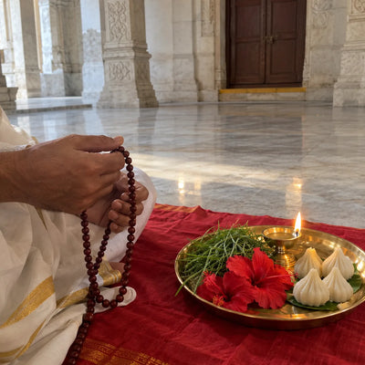 A red sandalwood mala beside durva grass, hibiscus, modaks and a ghee lamp
