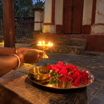Hands circling a brass aarti lamp beside red flowers, sindoor and a bowl of mustard oil