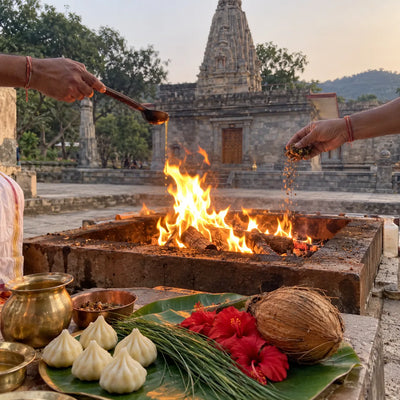 Two priests’ hands offering ghee into a havan fire with modaks and durva