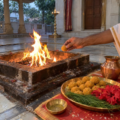 A laddoo offered into a havan fire beside a plate of laddoos and red hibiscus
