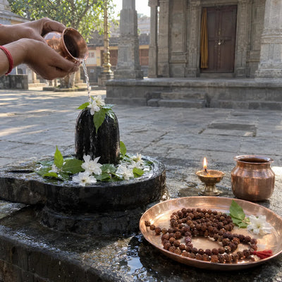A rudraksha mala on a copper plate beside a Shivling with bilva leaves and a small diya