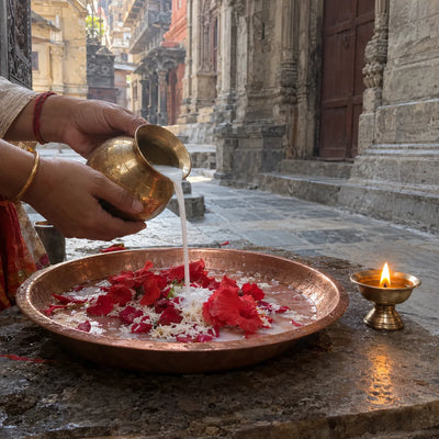 Hands pouring milk from a brass lota over red flowers and rice in a copper tray beside a ghee lamp