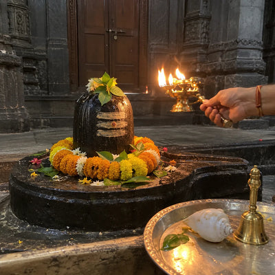 A multi-wick brass lamp waved before a garlanded black stone Shivling