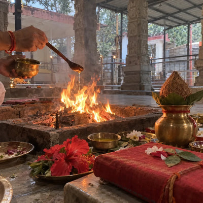 Hands offering ghee into a havan fire beside red hibiscus, a kalash with coconut and a wrapped pothi