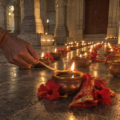 Rows of lit oil jyoti kalash lamps, one being lit in the foreground, with red hibiscus beside