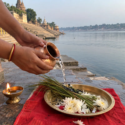 Hands pouring water with black sesame onto kusha grass beside white flowers and a lit diya