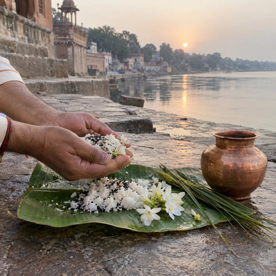 Hands offering cooked rice, black sesame and white flowers on a banana leaf beside a copper lota