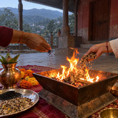 Hands offering black sesame and samagri into a havan fire beside a brass kalash and marigolds