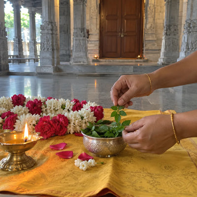 Fresh tulsi leaves in a silver bowl beside a rose and jasmine garland and a ghee diya