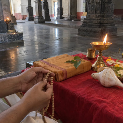 Hands holding a tulsi mala beside a wrapped prayer book, a brass lamp and a conch on red cloth