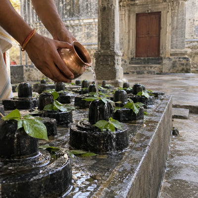 Water poured from a copper lota over rows of small black stone lingams with bilva