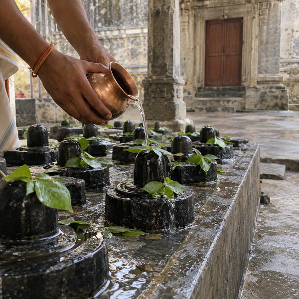 Water poured from a copper lota over rows of small black stone lingams with bilva