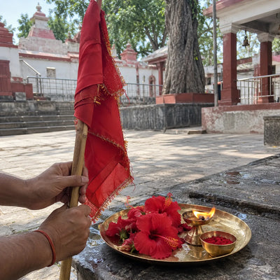 Hands holding a folded red flag beside red hibiscus, sindoor and a ghee lamp on a brass plate