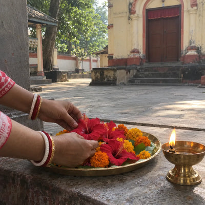 Hands placing red hibiscus and marigold flowers on a brass plate beside a lit ghee lamp