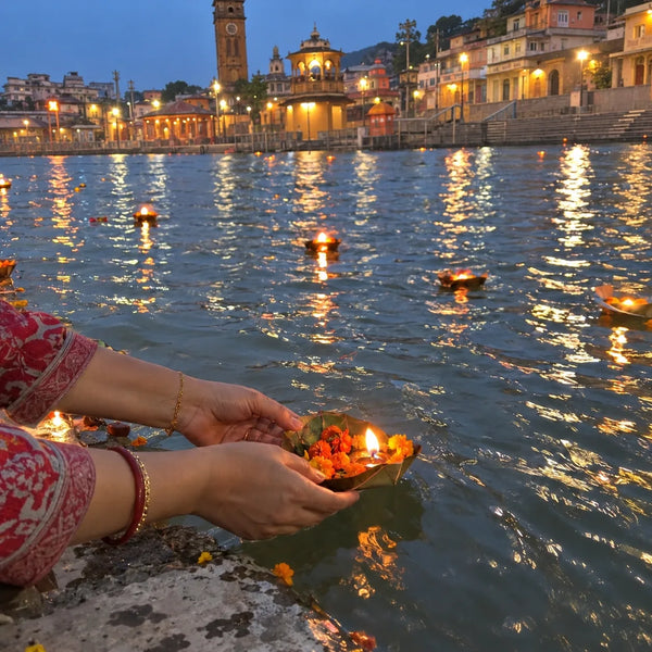 Hands releasing a flower-and-diya leaf cup onto the Ganga at dusk