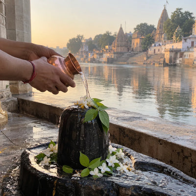 Hands pouring water from a copper lota onto a black stone lingam with bilva leaves