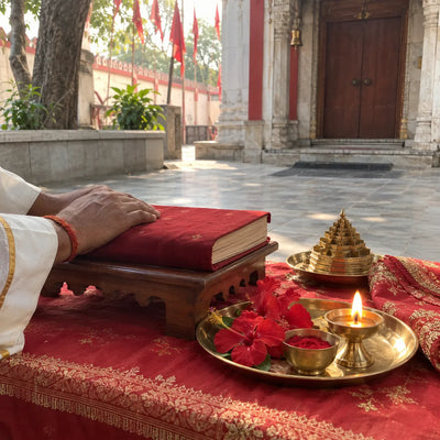 A cloth-bound scripture beside a brass Shree Yantra, red hibiscus, kumkum and a ghee diya