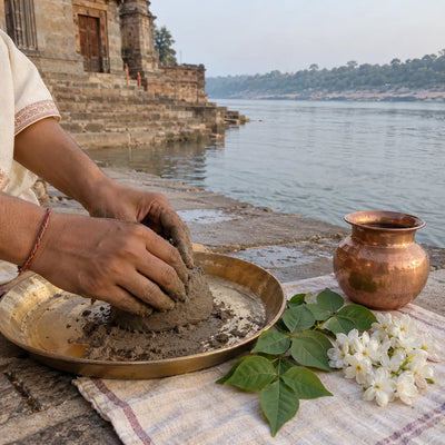 Hands shaping river clay on a brass plate beside bilva leaves, white flowers and a copper lota