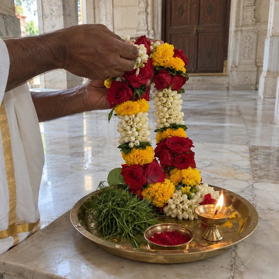 Hands holding a fresh garland of roses, marigolds and jasmine above a brass plate