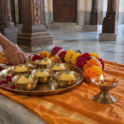 A brass rajbhog thali with tulsi leaves beside a marigold garland and a ghee diya on saffron cloth