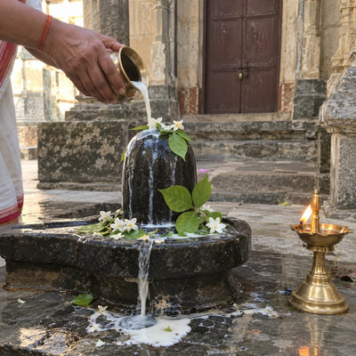 Hands pouring milk over a black stone lingam with bilva leaves, white flowers and a brass lamp
